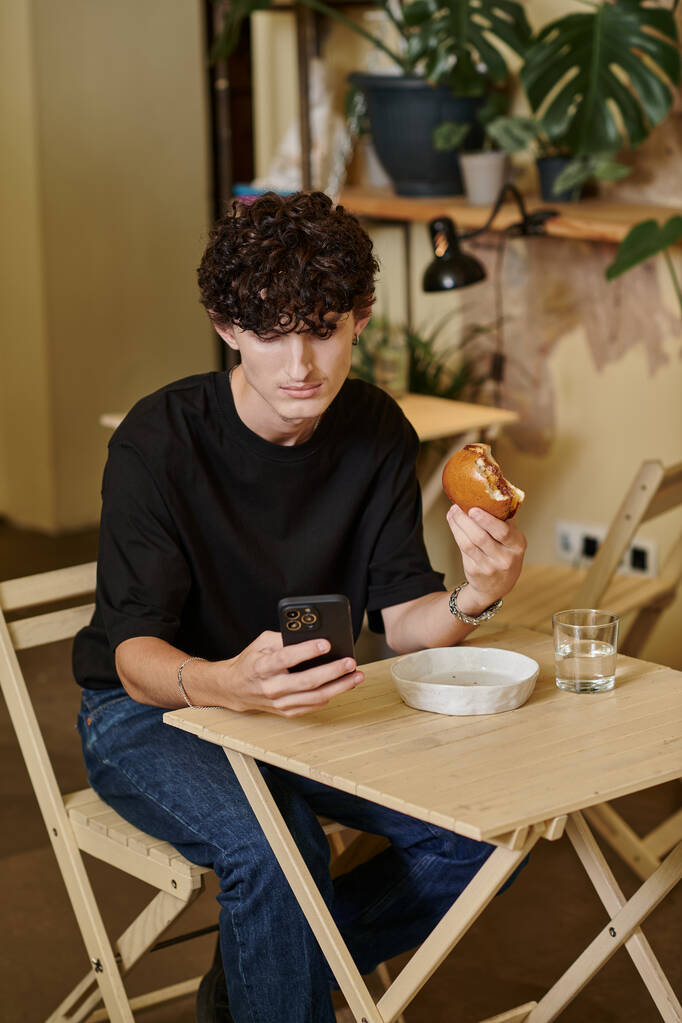 joven feliz y rizado sosteniendo a base de plantas de tofu hamburguesa mensajes de texto en el teléfono inteligente en la cafetería vegana - Foto, Imagen