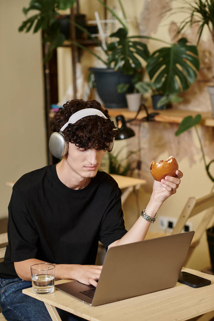 giovane uomo in cuffie wireless utilizzando il computer portatile e godendo di hamburger di tofu in caffè vegan, a base di piante - Foto, immagini