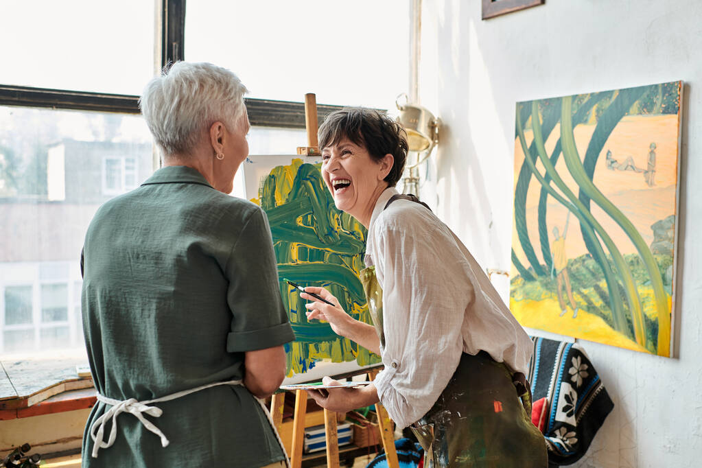 взволнованная зрелая женщина смеется возле мольберта и подруги в мастерской ремесла, творческое хобби - Фото, изображение