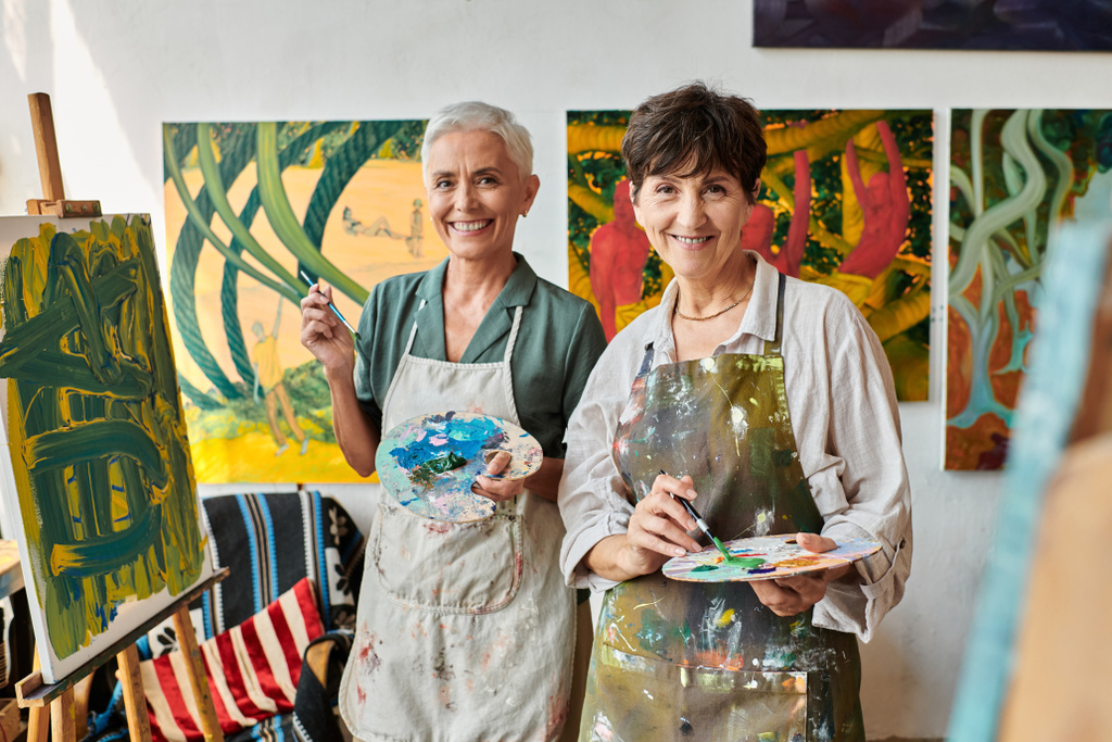 joyful mature female friends holding palettes and smile at camera in art studio, artistic hobby - Fotografie, Obrázek