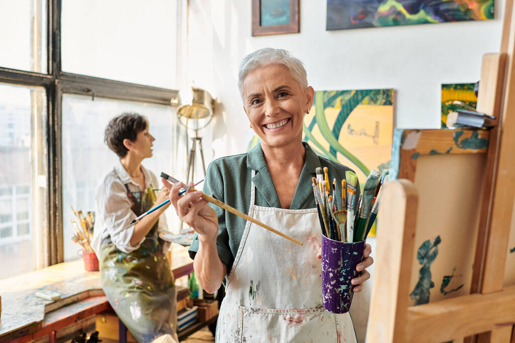 joyful mature woman with set of paintbrushes near female friend in art workshop, creative hobby - Photo, Image