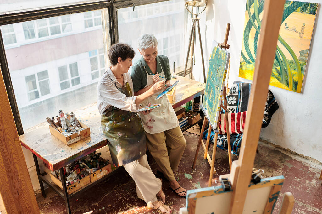 szczęśliwy w średnim wieku kobiety mieszanie kolorów na paletach w pobliżu sztalugi w studio sztuki, kreatywne hobby - Zdjęcie, obraz