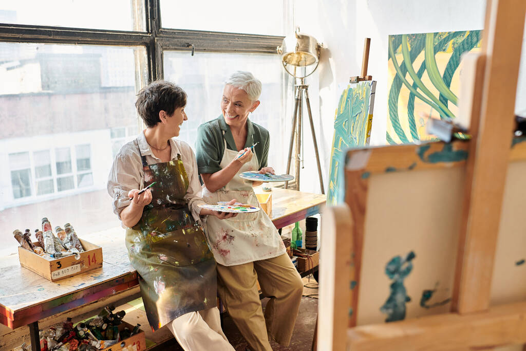 Reife Künstlerinnen mit Paletten und Pinseln unterhalten sich in Bastelwerkstatt nahe der Staffelei - Foto, Bild