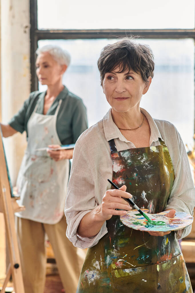 Gülümseyen olgun bir kadın renk paleti tutuyor ve sanat atölyesinde yüksek lisans yaparken başka tarafa bakıyor. - Fotoğraf, Görsel