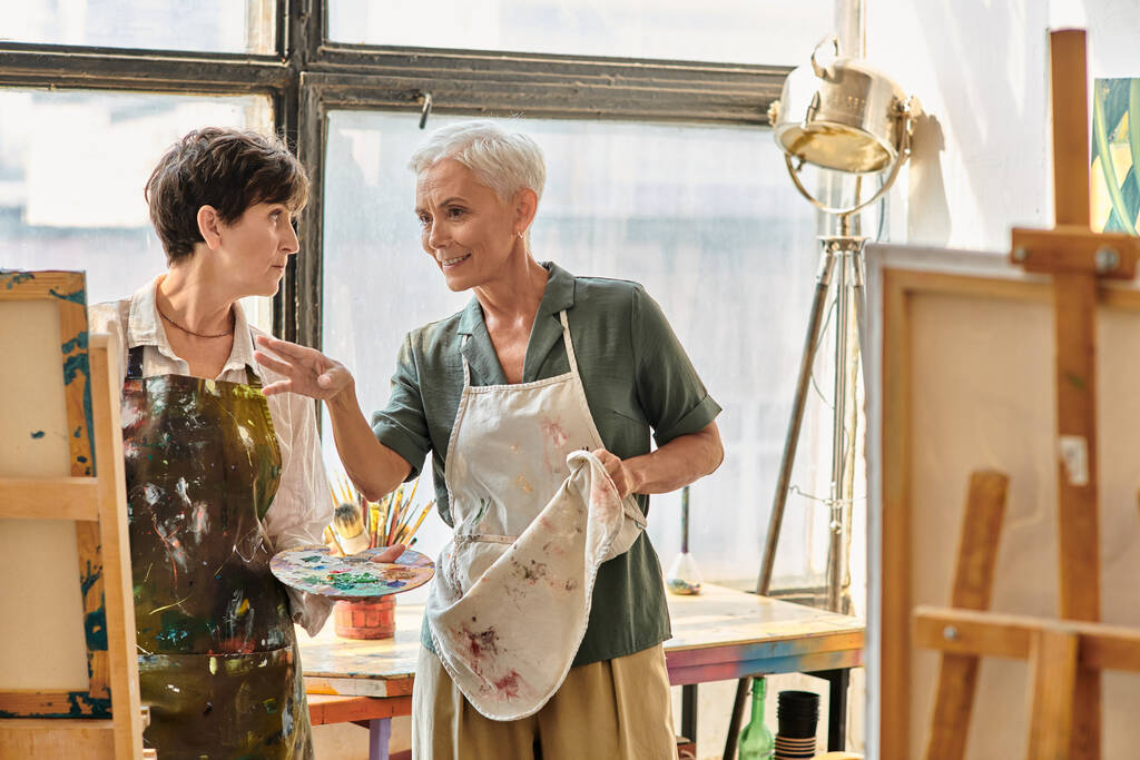 pozytywna artystka rozmawia z dojrzałą kobietą w fartuchu w pobliżu sztalugi podczas kursu mistrzowskiego w pracowni artystycznej - Zdjęcie, obraz