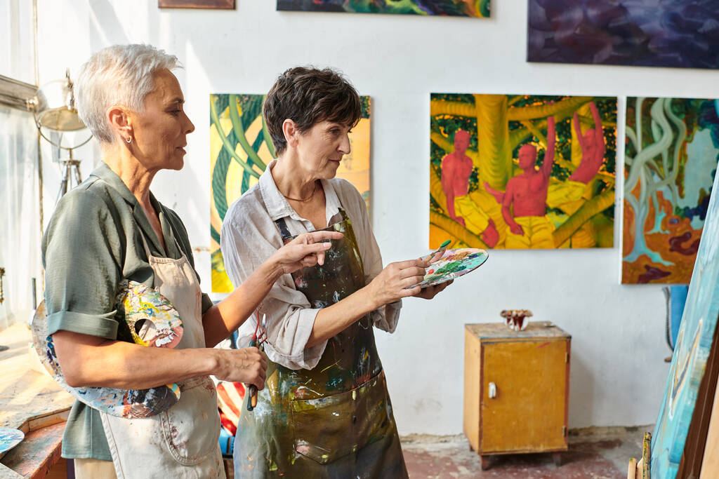 qualifizierte Künstlerin zeigt bei Meisterkurs im Atelier auf Staffelei nahe reifer Frau mit Palette - Foto, Bild