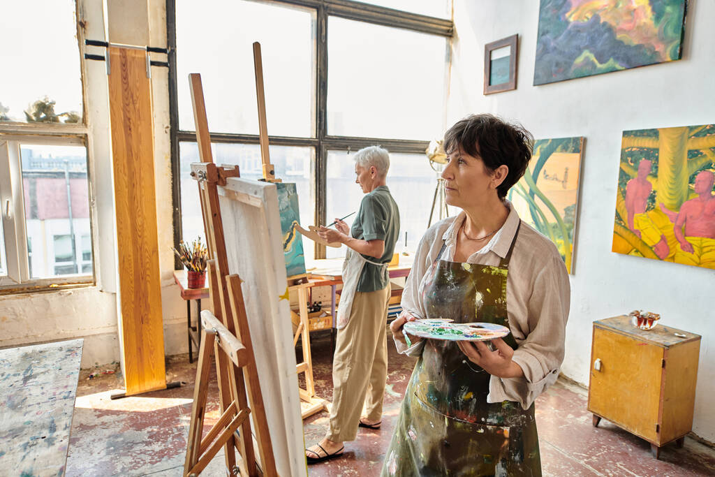 thoughtful mature woman in apron looking at easel near female friend in modern spacious art workshop - Photo, Image