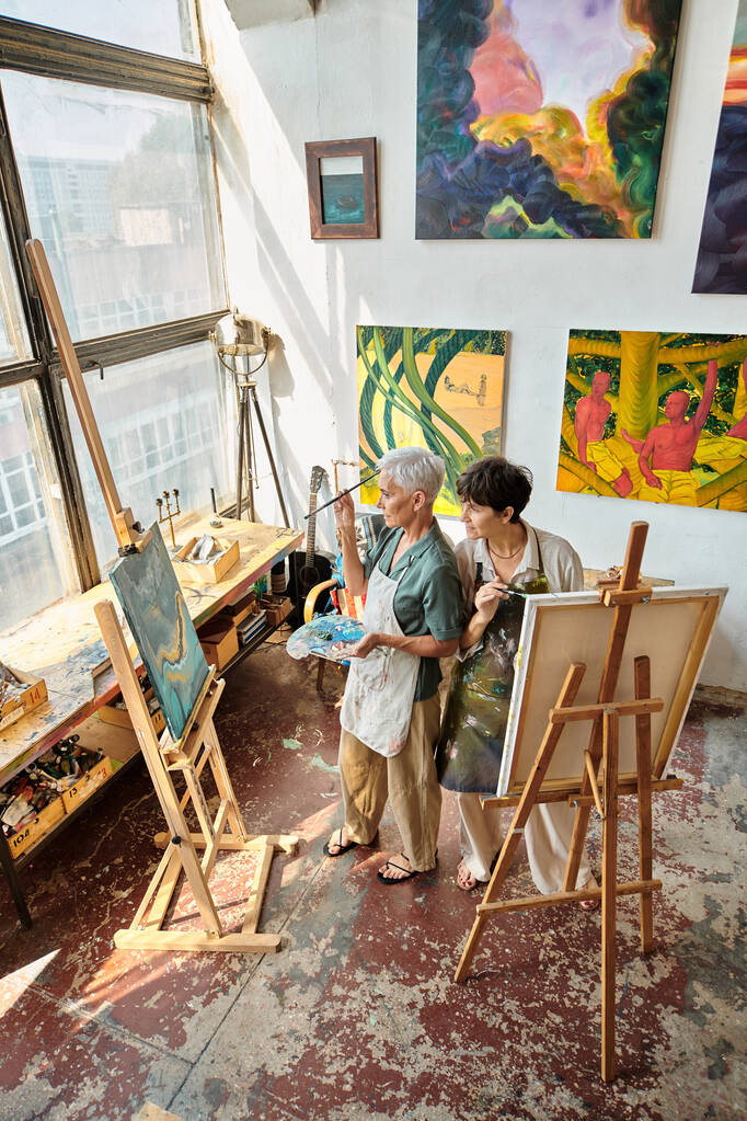 high angle view of mature female friends in aprons painting on easels in modern spacious workshop - Photo, Image