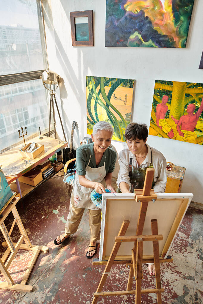 высокоугольный вид улыбающихся женщин-художниц в аппронах, глядящих на эспрессо в современной просторной мастерской - Фото, изображение