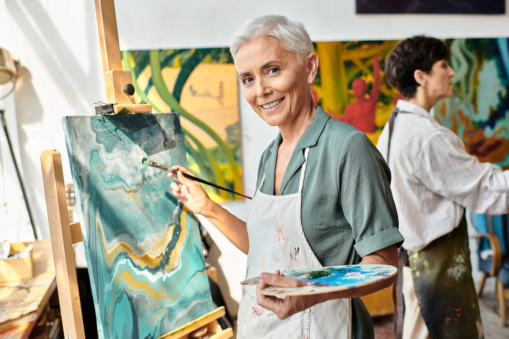 lächelnde reife Frau, die auf Staffelei malt und in der Kunstwerkstatt in die Kamera lächelt, kreativer Prozess - Foto, Bild