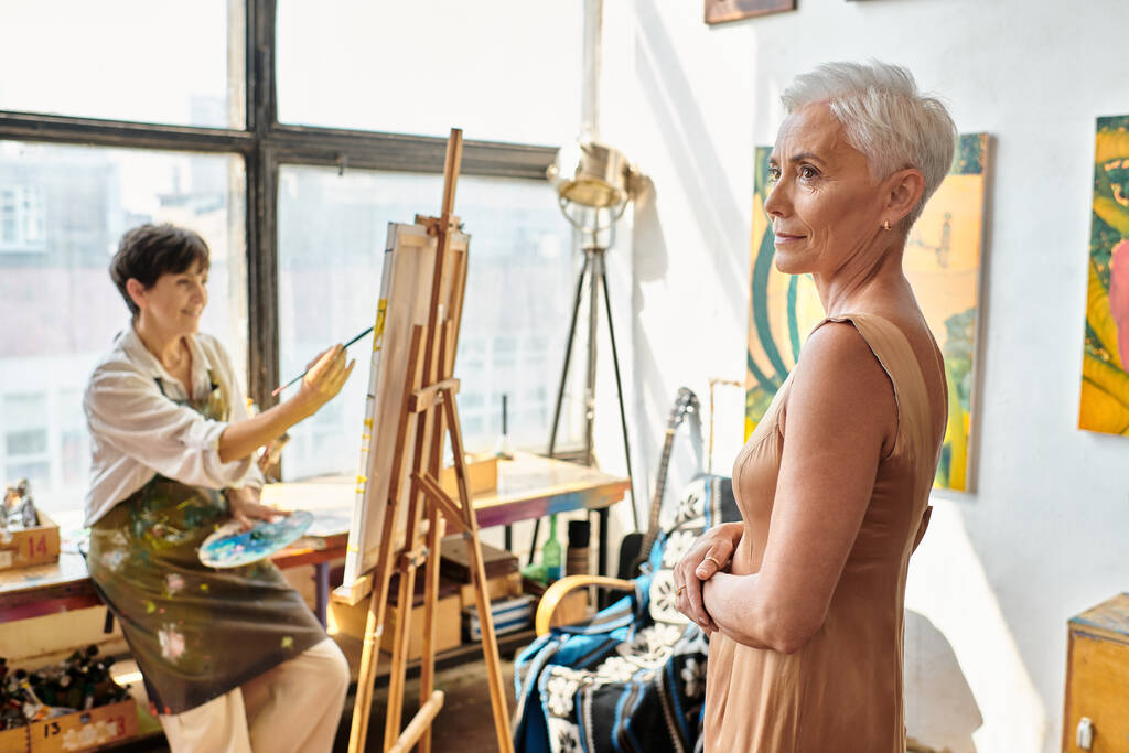 elegantes Modell mittleren Alters posiert in der Nähe weiblicher Künstler Malerei in der Werkstatt, kreativer Prozess - Foto, Bild