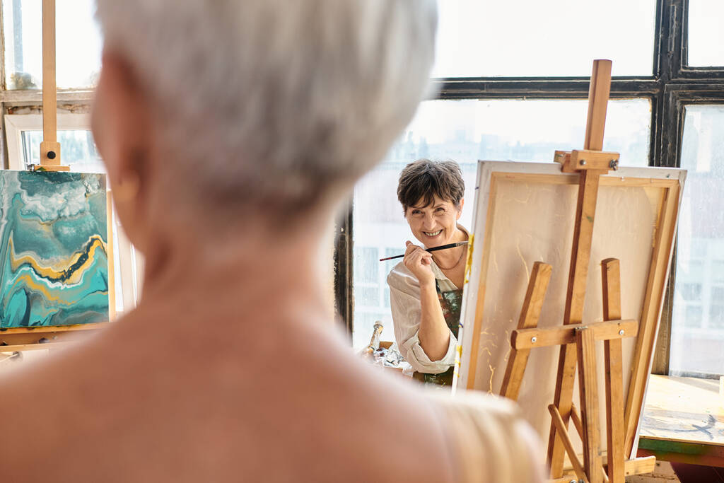 joyful female artist talking to blurred mature model posing in craft workshop, creative process - Photo, Image