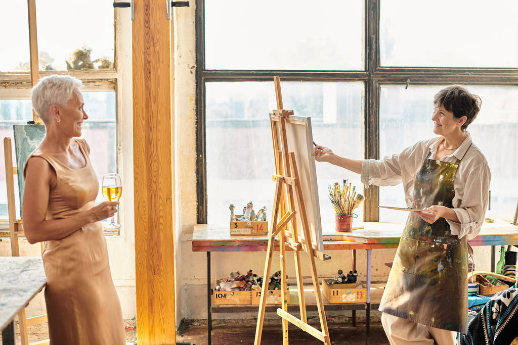 アートスタジオでワイングラスとポーズエレガントな女性の友人を描く幸せな成熟したアーティスト,創造性 - 写真・画像