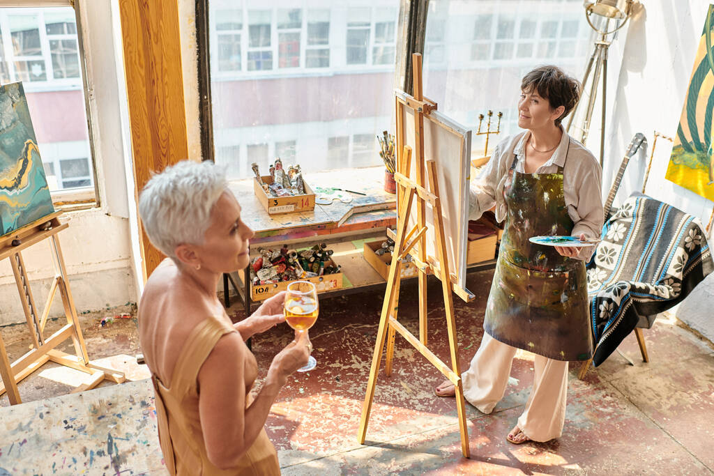 εμπνευσμένη ώριμη καλλιτέχνης ζωγραφική χαριτωμένο μοντέλο με γυαλί κρασιού στο εργαστήριο τέχνης, δημιουργική διαδικασία - Φωτογραφία, εικόνα