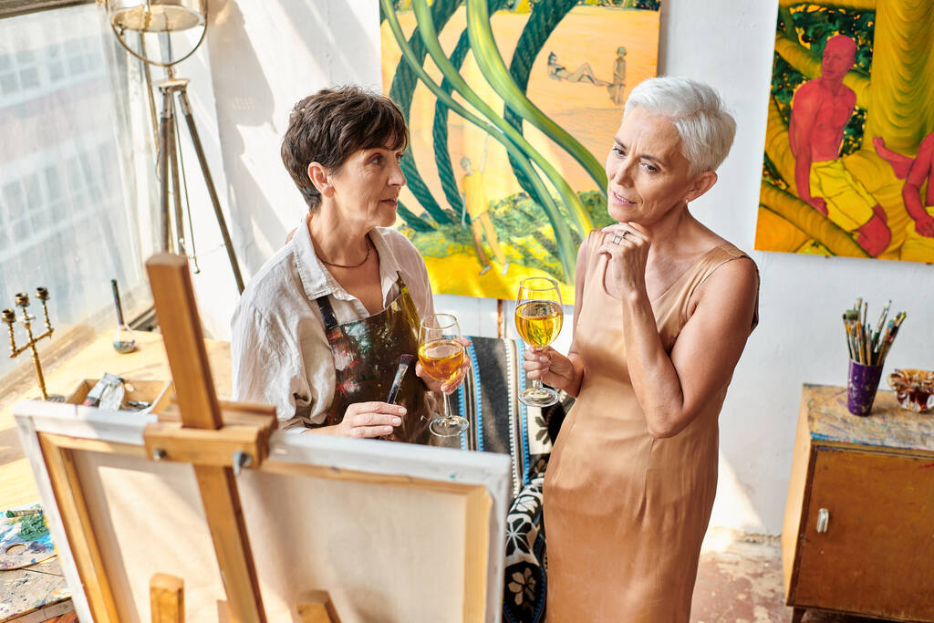 Şarap kadehiyle düşünceli olgun bir model stüdyoda profesyonel bir kadın sanatçının yanında resim sehpasına bakıyor. - Fotoğraf, Görsel