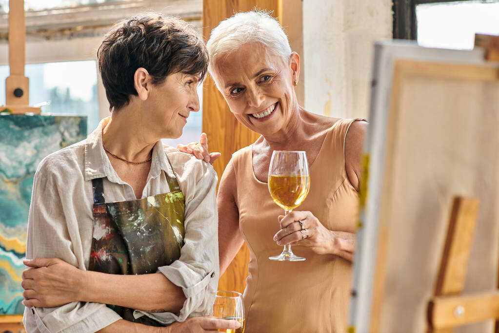 весёлая зрелая модель с бокалом вина, смотрящая на мольберт рядом с художницей в мастерской - Фото, изображение