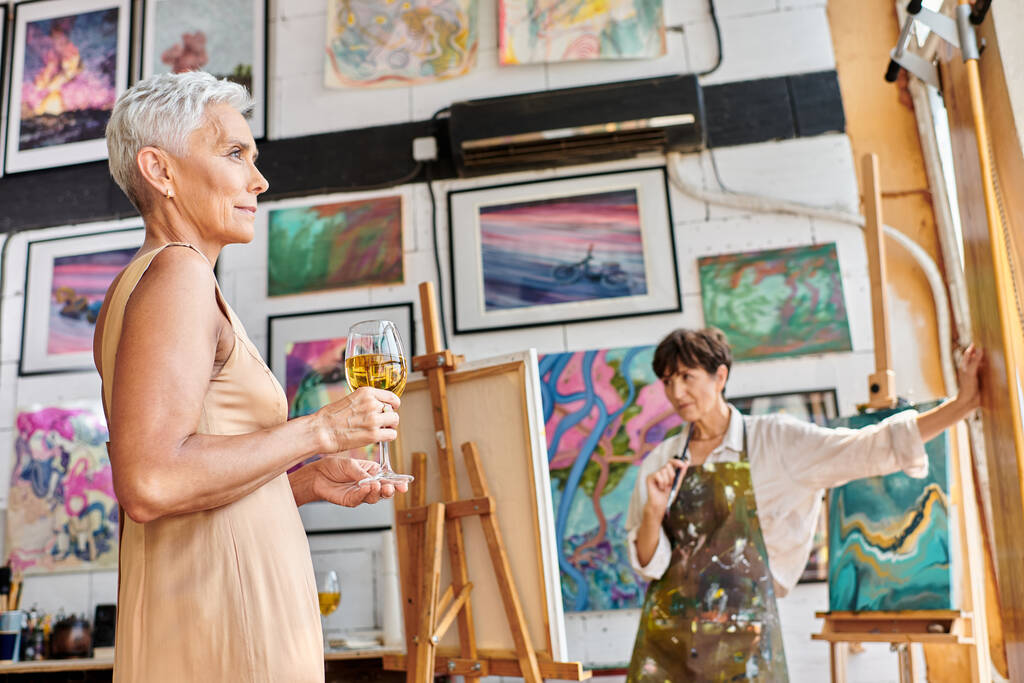 elegancki dojrzały model z kieliszkami do wina pozowanie w pobliżu kobiety malarz w studio sztuki, proces twórczy - Zdjęcie, obraz