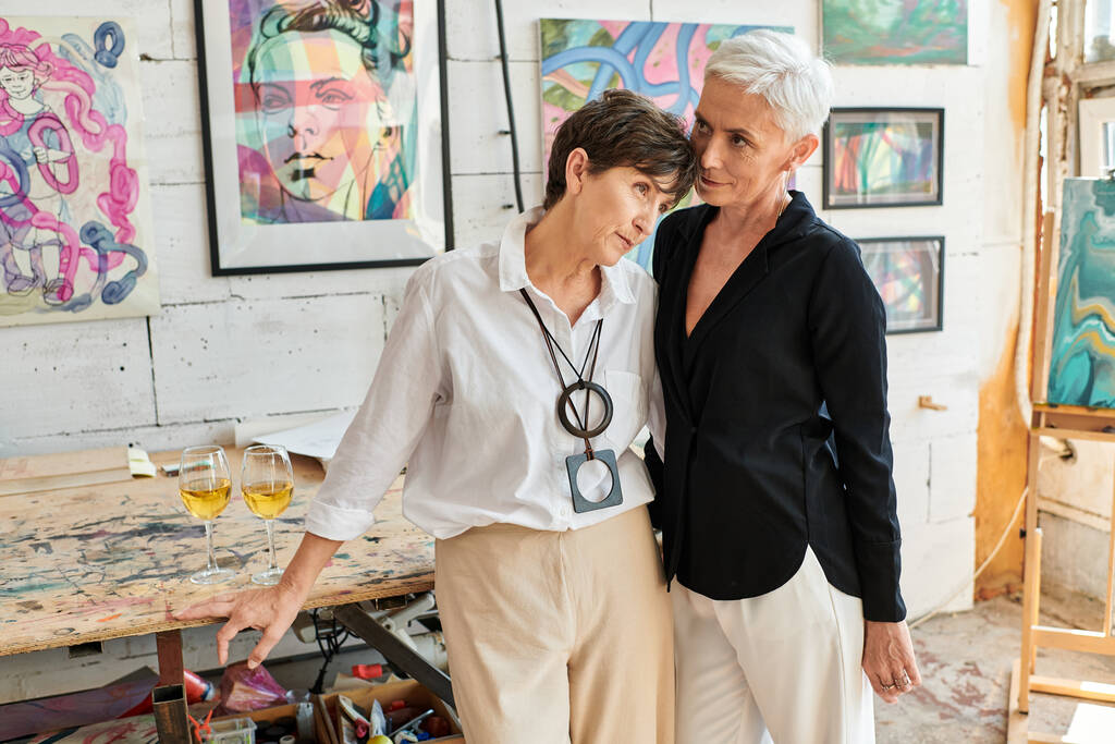 stylový dospělý lesbický pár stojící v blízkosti barevných obrazů v moderní výtvarné dílně, spolu - Fotografie, Obrázek