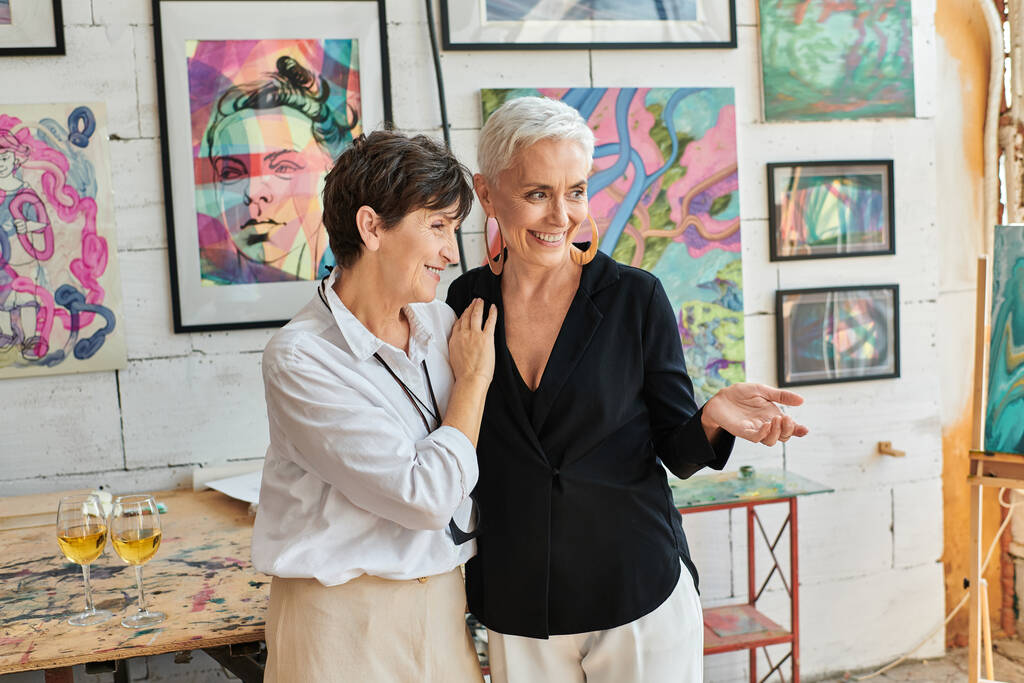 модная художница, указывающая рукой на радостного лесбийского партнера и картины в художественной мастерской - Фото, изображение