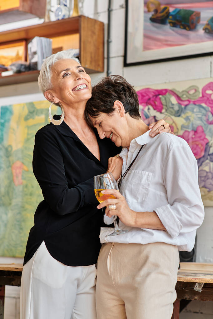 ενθουσιασμένοι μεσήλικη γυναίκα αγκαλιάζει λεσβία σύντροφο με γυαλί κρασιού στο εργαστήριο σύγχρονης τέχνης - Φωτογραφία, εικόνα