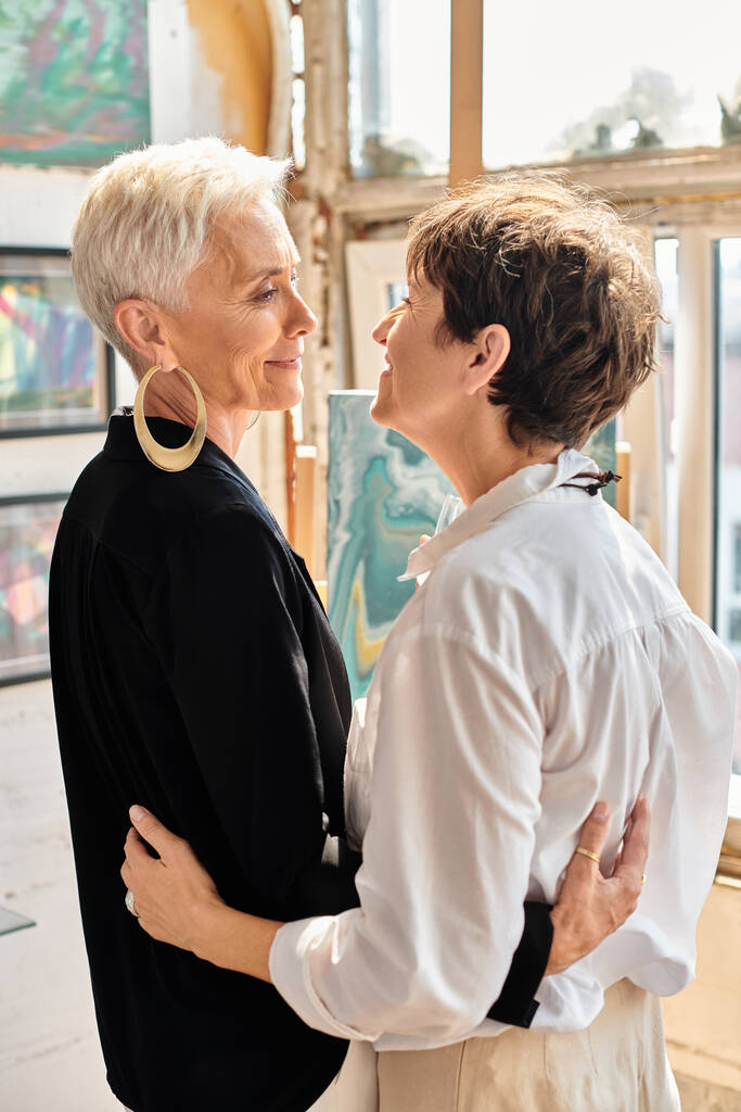 веселые и элегантные зрелые лесбийские художники, смотрящие друг на друга в студии современного искусства - Фото, изображение