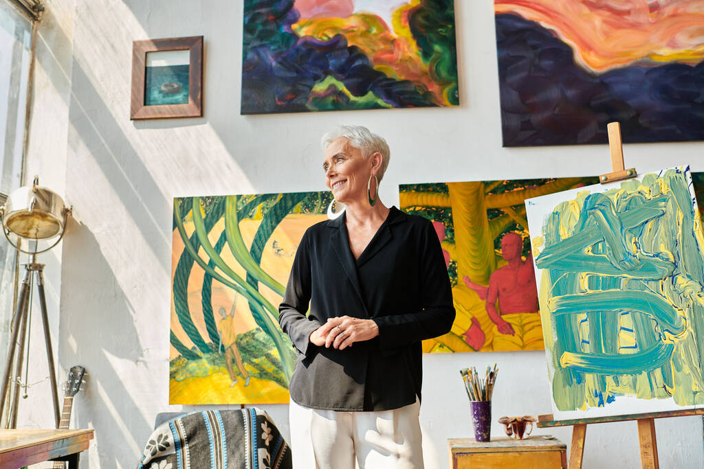 stijlvolle vrouwelijke kunstenaar van middelbare leeftijd glimlachend en wegkijkend in de buurt van kleurrijke schilderijen in kunststudio - Foto, afbeelding