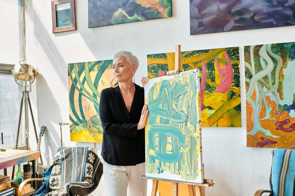 harkittuja ja trendikäs naisartisti seisoo lähellä maalausteline ja värikkäitä luovia maalauksia studiossa - Valokuva, kuva