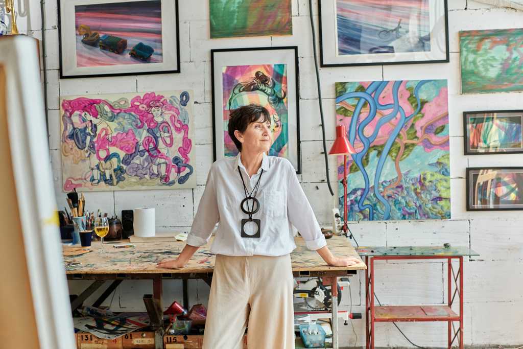 przemyślany dojrzała kobieta artysta stojąc i patrząc daleko w studio sztuki z kolorowymi obrazami - Zdjęcie, obraz