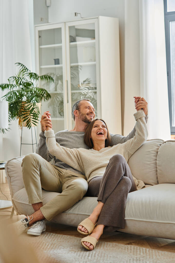 excited child-free couple holding hands and laughing on cozy couch in living room, fun and leisure - Photo, Image