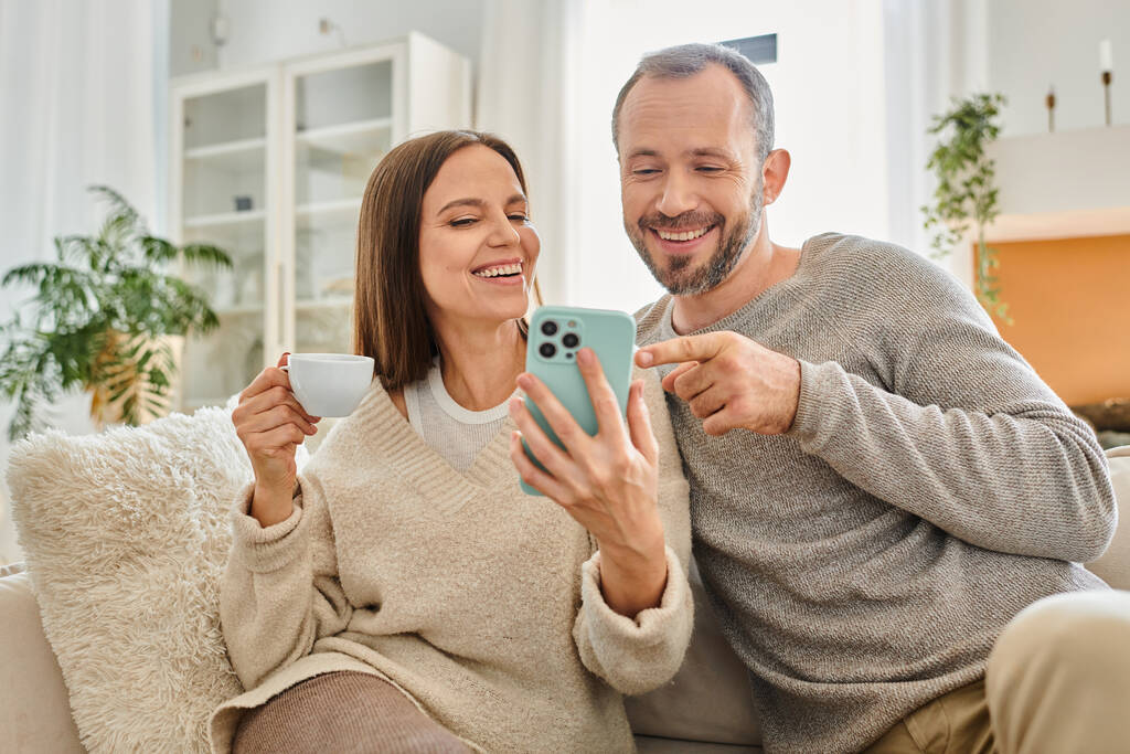 hombre alegre apuntando al teléfono inteligente cerca de la esposa con taza de café en el sofá en casa, estilo de vida libre de niños - Foto, Imagen