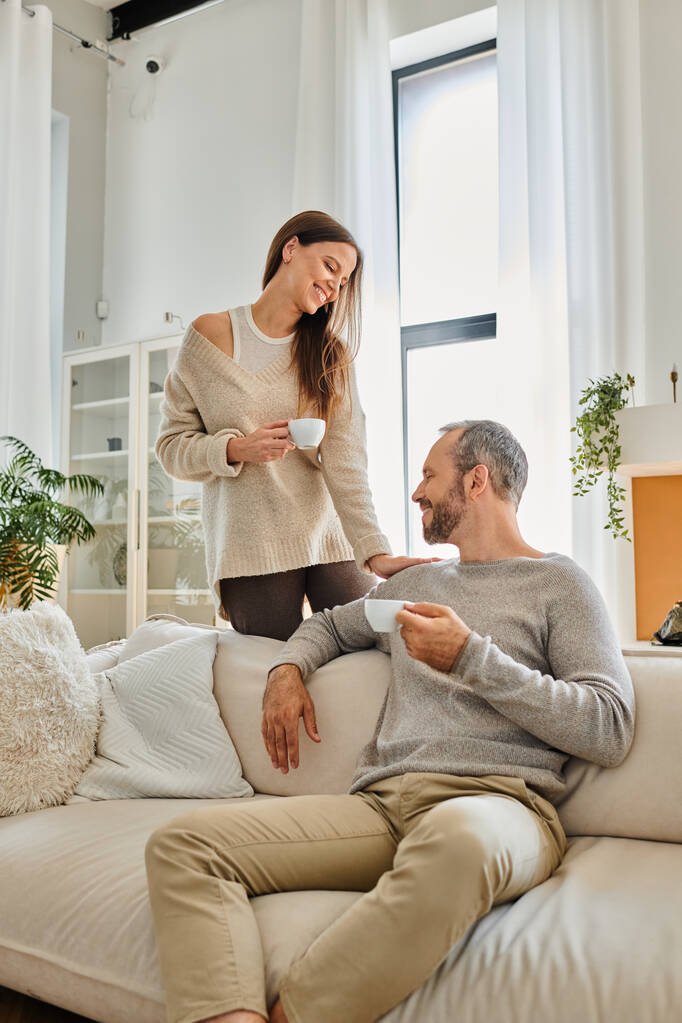 fröhliches kinderfreies Paar trinkt Morgenkaffee im modernen Wohnzimmer der gemütlichen Wohnung, Gelassenheit - Foto, Bild