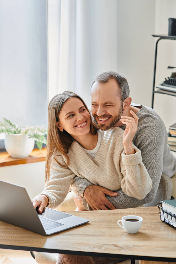 збуджений чоловік обіймає щасливу дружину, працюючи на ноутбуці вдома, догляд та підтримка бездітної пари - Фото, зображення