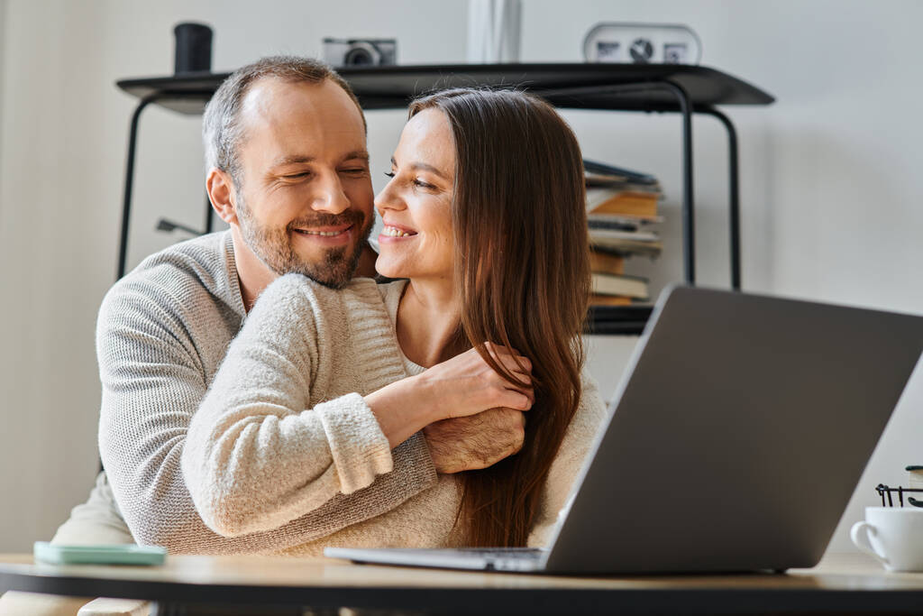 glücklicher Ehemann umarmt fröhliche Ehefrau, die zu Hause am Laptop arbeitet, Pflege und Unterstützung von kinderfreien Paaren - Foto, Bild