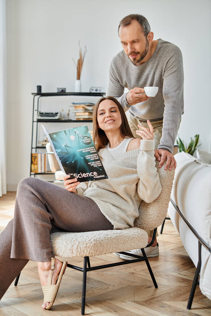 счастливая женщина, читающая научный журнал рядом с мужем с чашкой кофе на диване дома, жизнь без детей - Фото, изображение