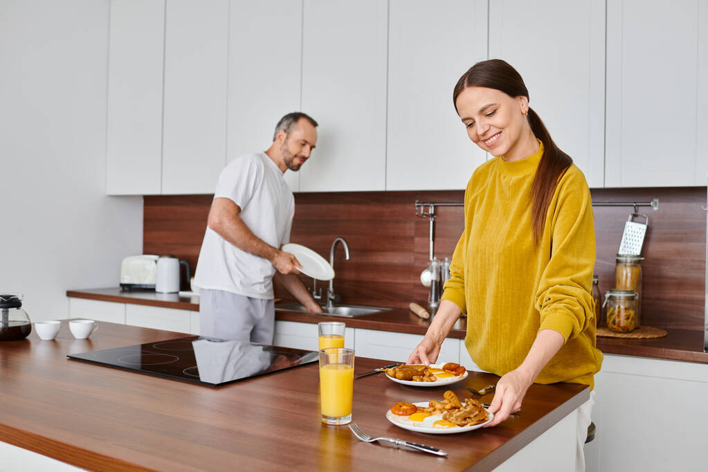 mujer sonriente que sirve sabroso desayuno mientras el marido lava los platos en la cocina, el cuidado y la armonía - Foto, imagen