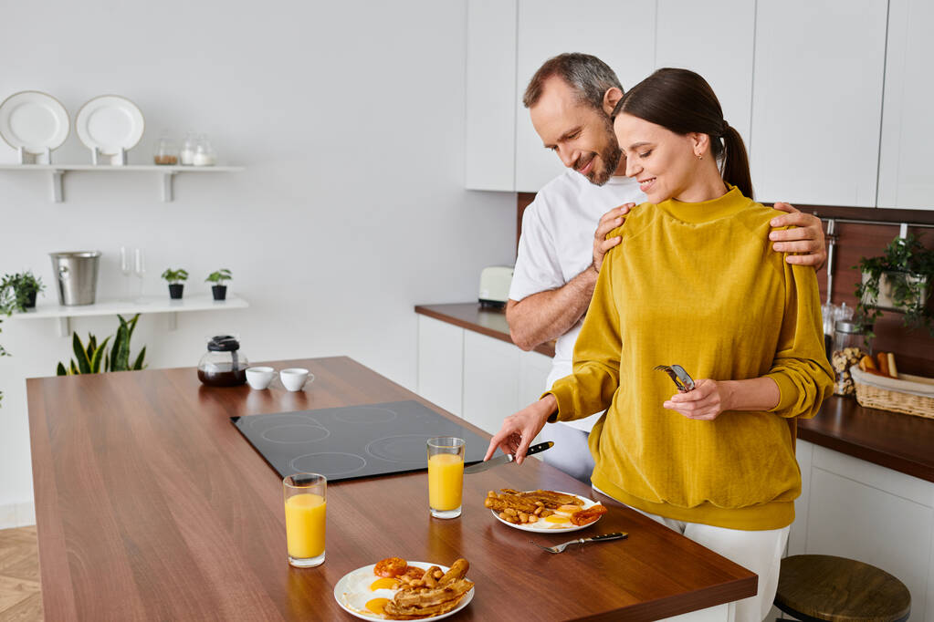 щасливий чоловік обіймає плечі дружини, подаючи смачний сніданок на кухні, вранці бездітна пара - Фото, зображення