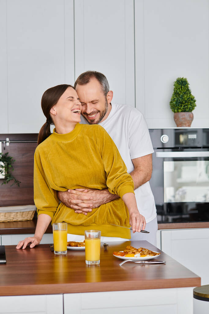 キッチンでおいしい朝食の近くで笑う妻を抱擁する幸せな男,子供のないカップルの朝 - 写真・画像
