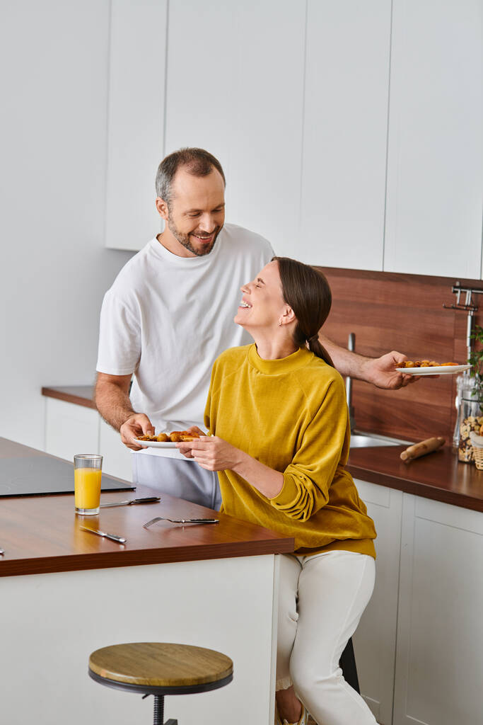 Улыбающийся мужчина, подающий вкусный завтрак рядом со смеющейся женой на кухне, утро свободной от детей пары - Фото, изображение
