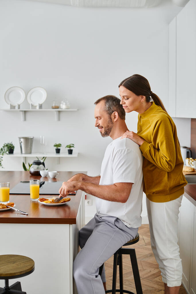 вид сбоку улыбающегося мужчины, наслаждающегося вкусным завтраком рядом с заботливой женой на кухне, бездетная пара - Фото, изображение
