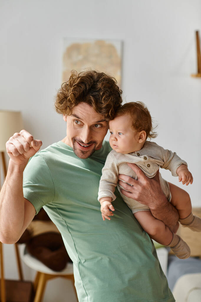 彼の幼い息子を抱きしめ,居心地の良い寝室で指で指を指差しているカーリー髪の父親 - 写真・画像