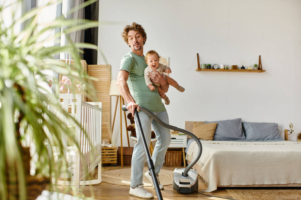 homem multitarefa trabalho doméstico e puericultura, encaracolado pai aspirando apartamento com menino nos braços - Foto, Imagem