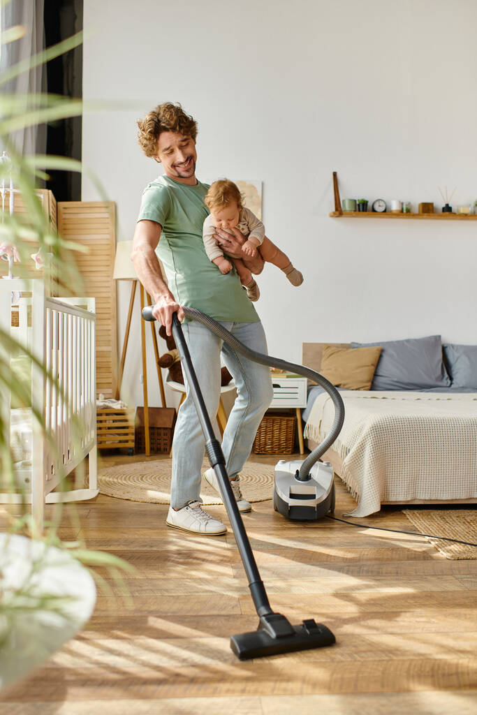 мужчина многозадачная работа по дому и уходу за детьми, улыбающийся отец пылесосит квартиру с младенцем на руках - Фото, изображение