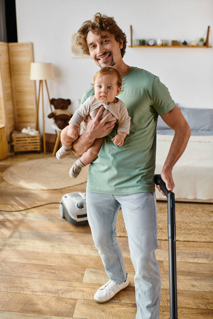 szczęśliwy człowiek wielozadaniowe prace domowe i opieka nad dziećmi, ojciec odkurzanie podłogi z drewna twardego z synem w ramionach - Zdjęcie, obraz
