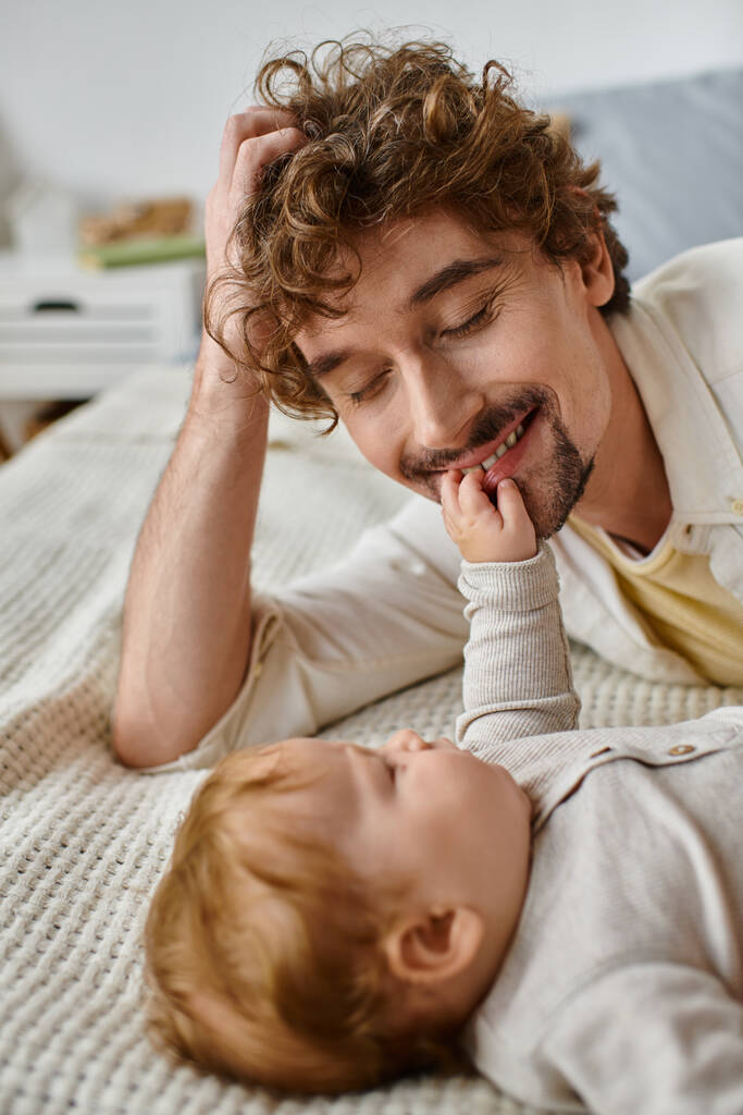 lindo bebé niño acostado en la cama y tocando la cara de su padre feliz barbudo, momentos preciosos - Foto, imagen