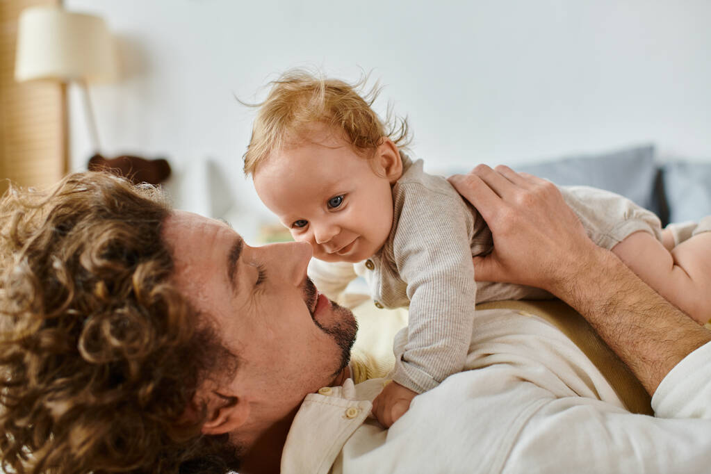 szczęśliwy człowiek z kręconymi włosami i brodą obejmujący swojego synka w sypialni, miłości i opieki - Zdjęcie, obraz