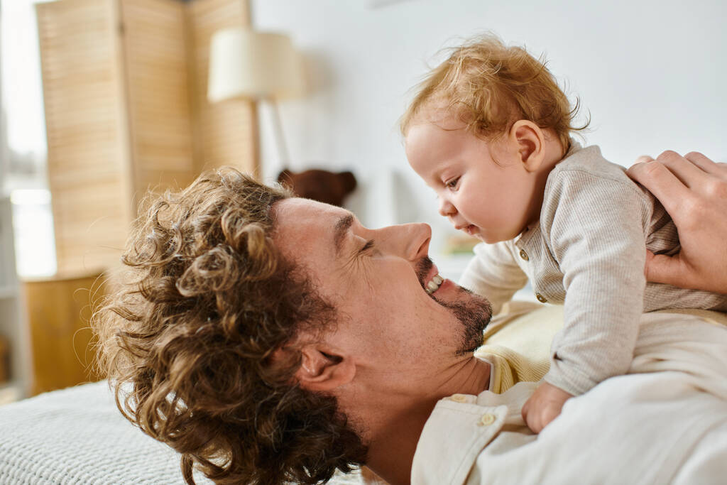 glücklicher Mann mit lockigem Haar und Bart mit Blick auf seinen kleinen Sohn im Schlafzimmer, Liebe und Fürsorge des Vaters - Foto, Bild