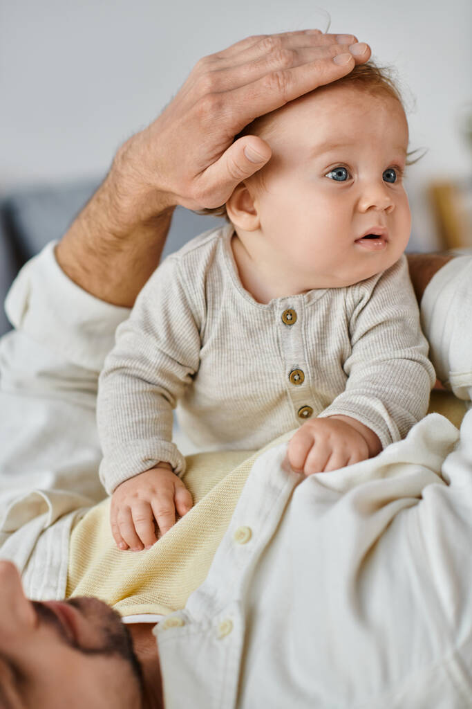 svobodný otec s kudrnatými vlasy a vousy hladící vlasy svého kojeneckého chlapce v ložnici, láska a péče - Fotografie, Obrázek