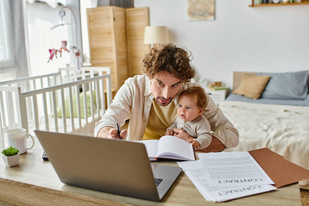 man holding in arms his infant son while taking notes and working from home, work-life balance - Photo, Image