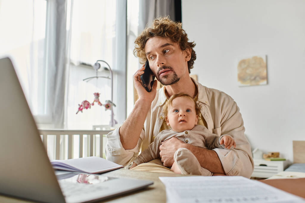 Vater hält kleinen Sohn beim Telefonieren auf Smartphone und arbeitet von zu Hause aus in der Nähe von Papieren und Laptop - Foto, Bild