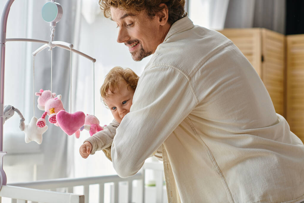 caring single father holding his infant son near baby mobile in nursery, fatherhood and love - Photo, Image